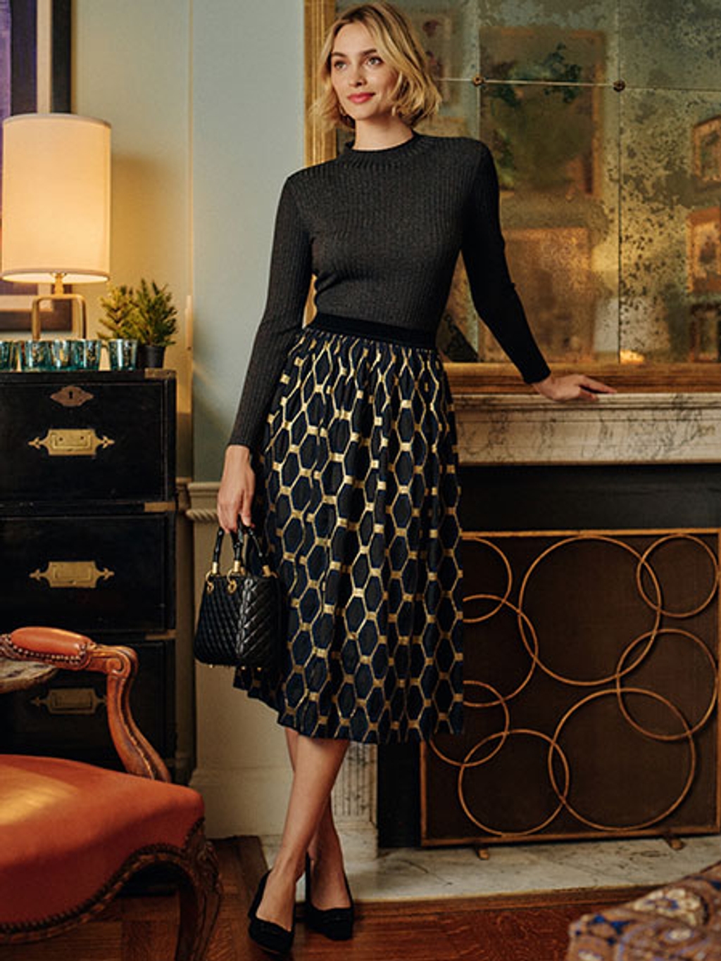 Model wearing rib-knit turtleneck, jacquard skirt, and heels in front of fireplace