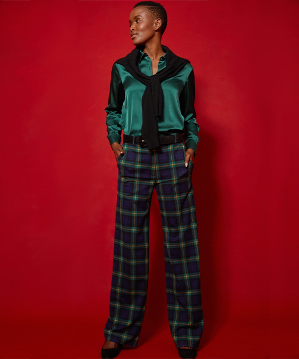 Model wearing tartan pants, satin button-down, cashmere sweater, leather belt, and suede pumps.
