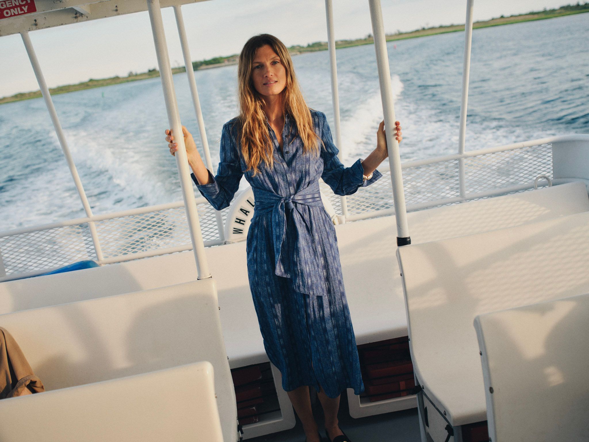 A woman in a blue patterned dress stands on a boat, holding onto rails, with the ocean and a wake behind her.