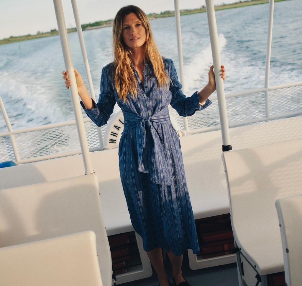 Woman in blue striped dress stands on a boat, looking at the camera, with water and distant land.