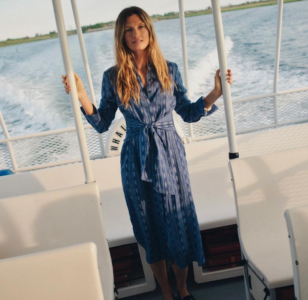 A woman in a blue patterned dress stands on a boat, holding onto rails, with the ocean and a wake behind her.