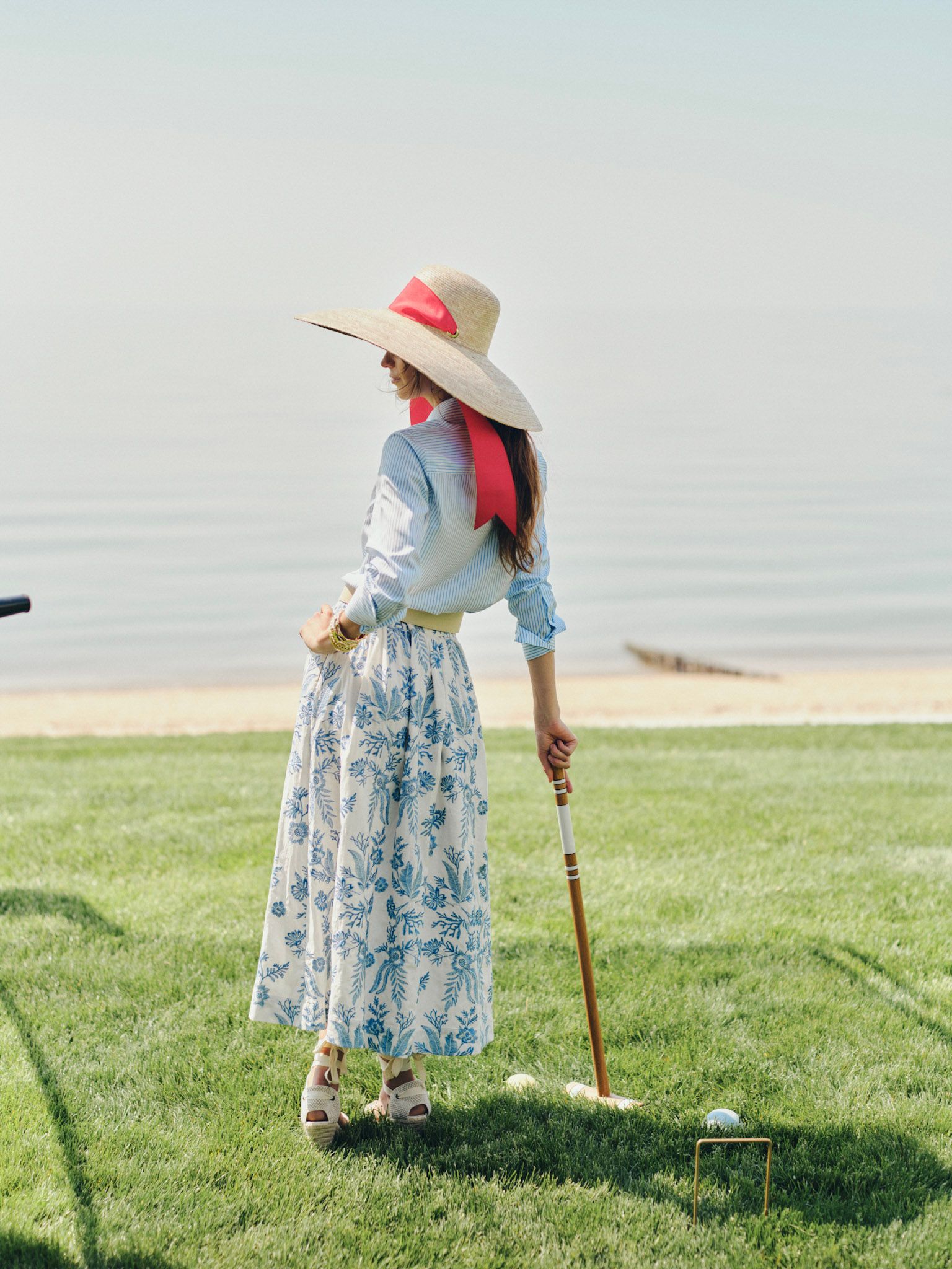 Woman in a straw hat with a red ribbon and blue floral skirt playing croquet on a lawn by the water.