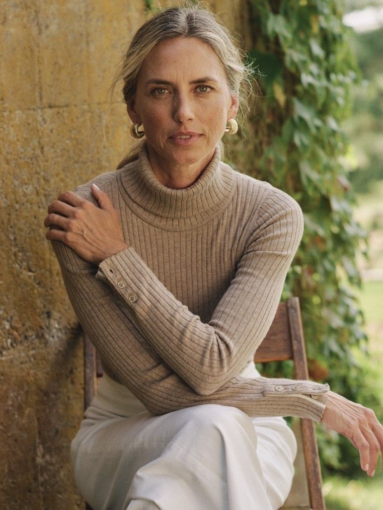 A woman in a tan ribbed turtleneck, light pants, and gold hoop earrings sits looking forward, with a stone wall and greenery in the background.
