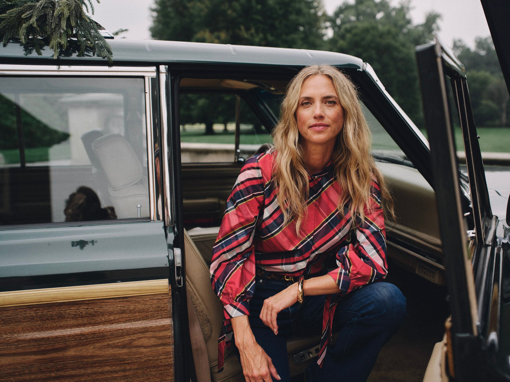 A blonde woman in a red plaid shirt and jeans sits in the open door of a vintage green wood-paneled station wagon with a Christmas tree on its roof.