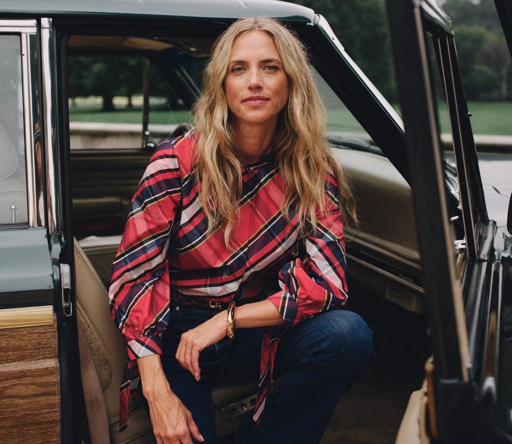 A blonde woman in a red plaid shirt and jeans sits in the open door of a vintage green wood-paneled station wagon with a Christmas tree on its roof.