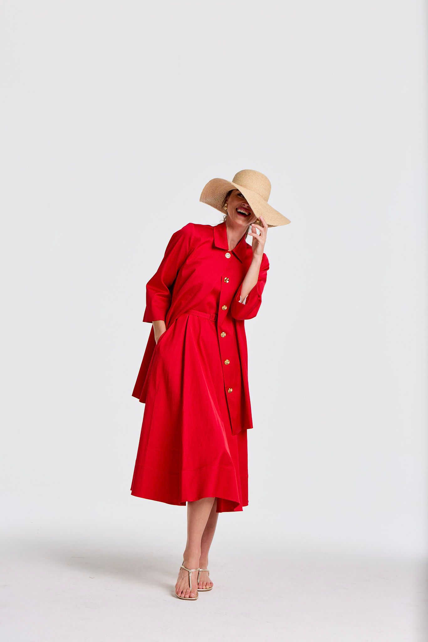 A woman in a red dress and straw hat laughs, covering her mouth with her hand.