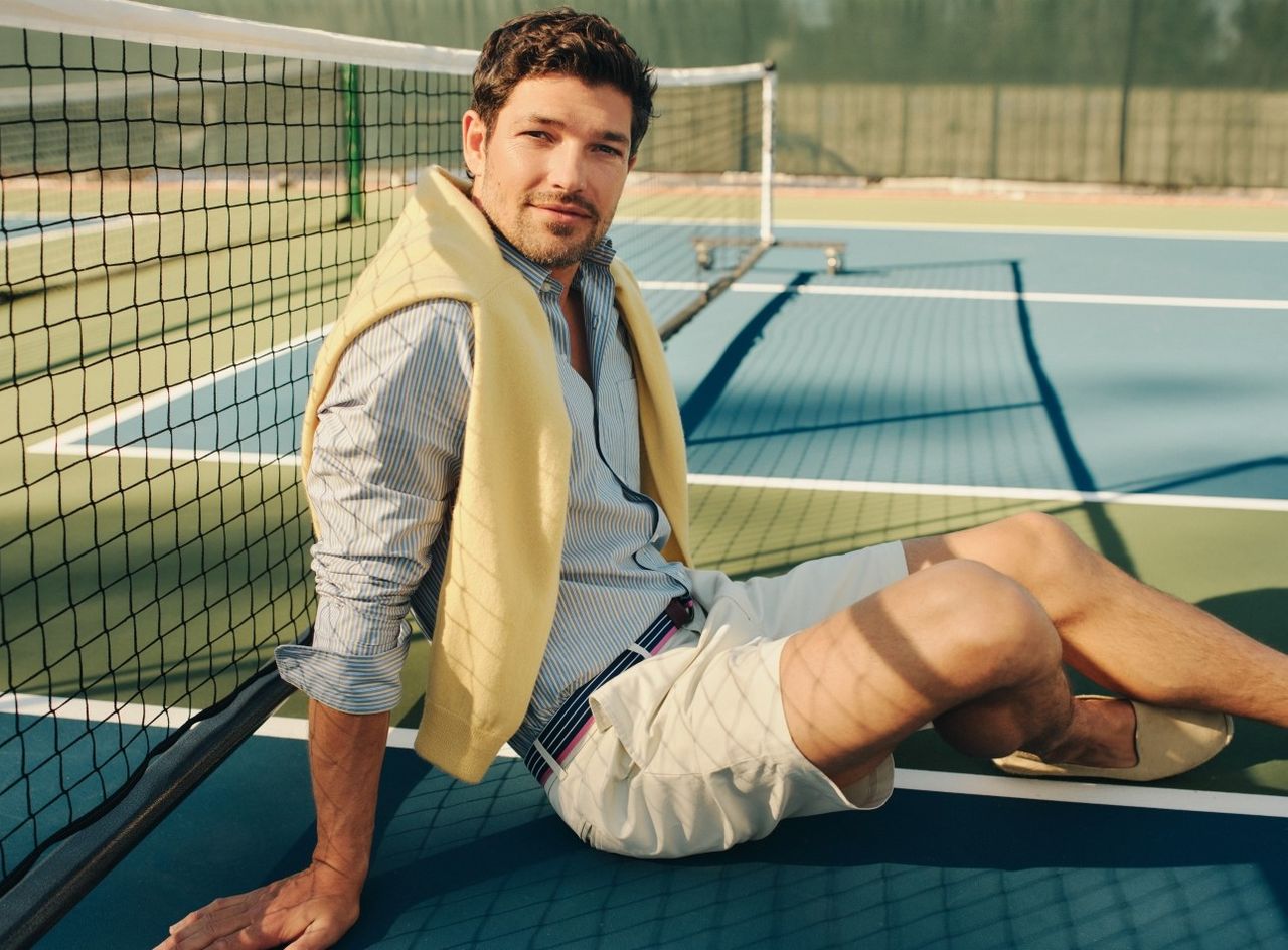 man on tennis court seated, wearing khaki chino short and blue stripe button down shirt with yellow sweater over shoulders