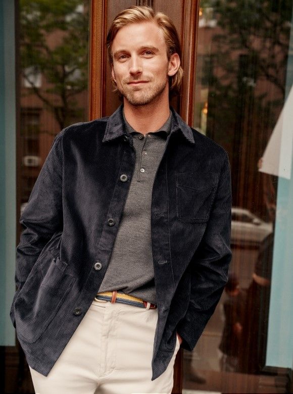 man leaning against door outside wearing navy suede jacket, gray knit polo and khaki trousers