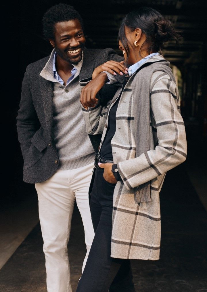 woman and man standing outside. Her wearing light gray plaid wool mid length coat. Him wearing light gray polo sweater layered underneath a gray herringbone sport coat