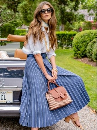 woman seated on back of vintage looking car in navy pleated skirt and white linen shirt