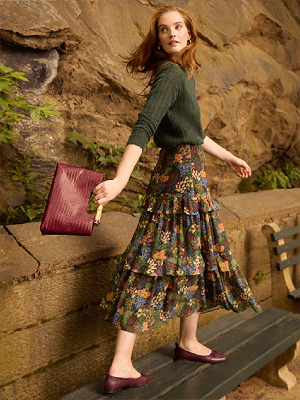 Woman looking back over her shoulder holding a red handbag, wearing a patterned skirt.