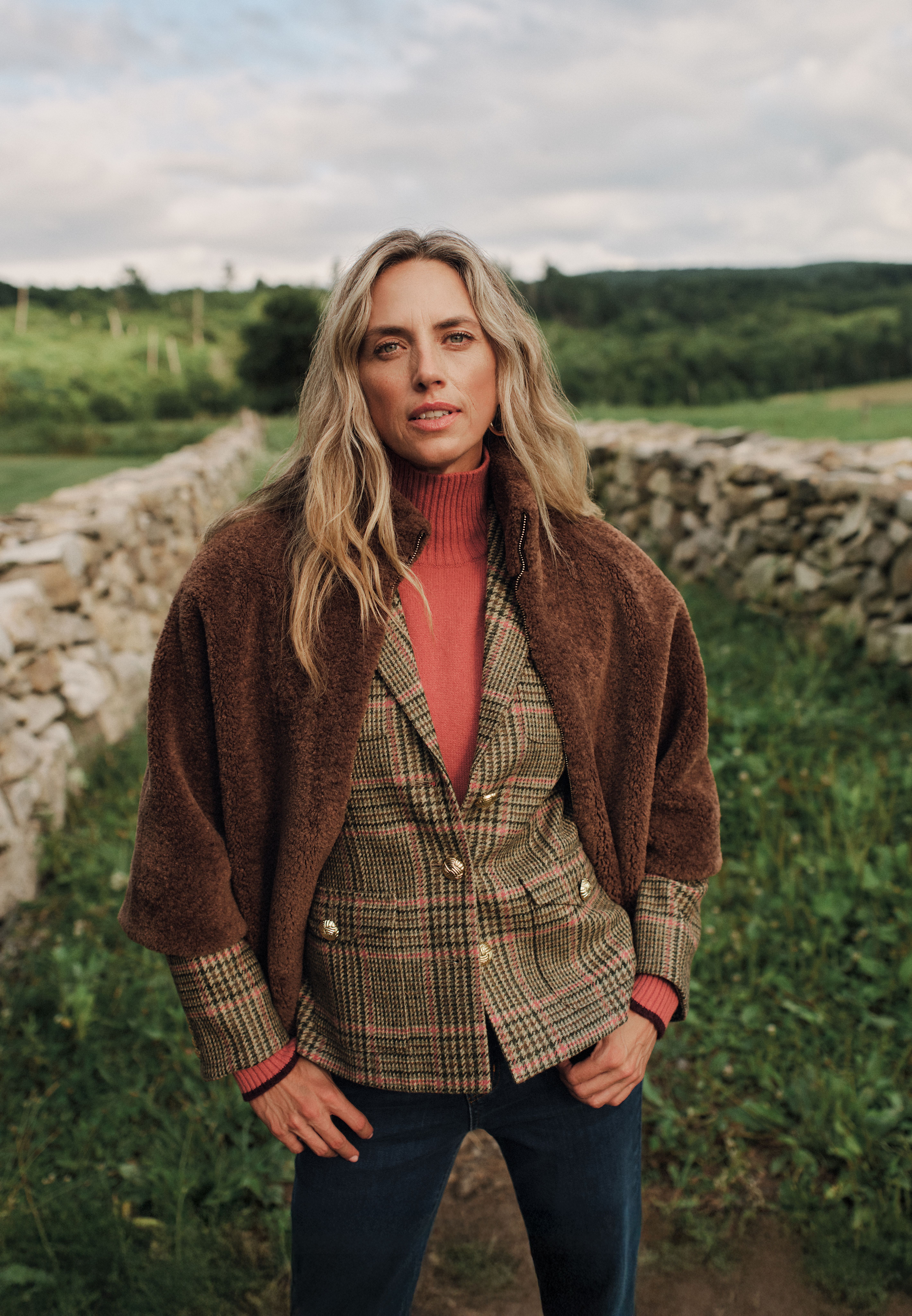A woman with long blonde hair wears a brown faux fur jacket, pink turtleneck, and plaid blazer outdoors with a stone wall behind her.