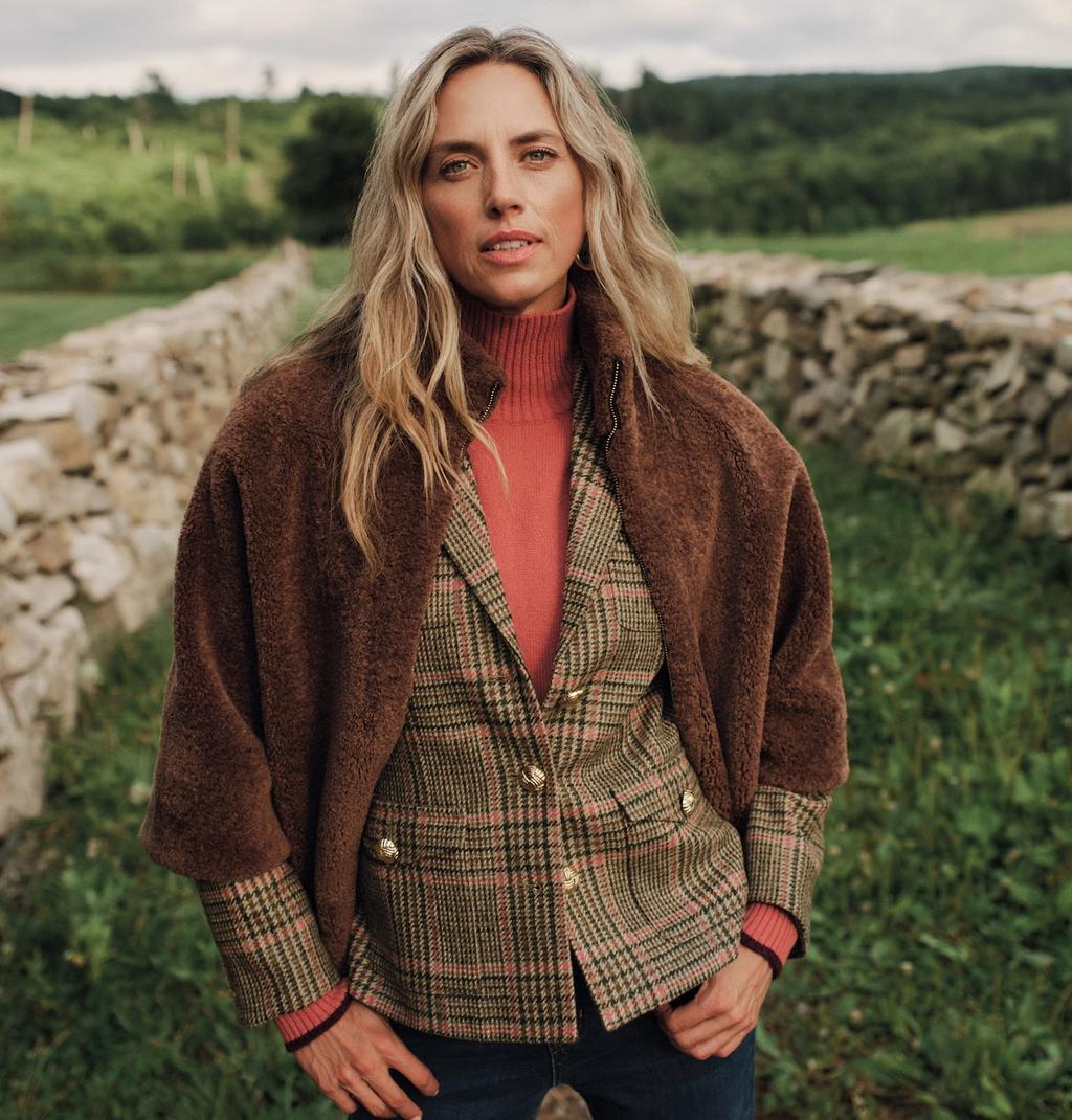 A woman with long blonde hair wears a brown faux fur jacket, pink turtleneck, and plaid blazer outdoors with a stone wall behind her.