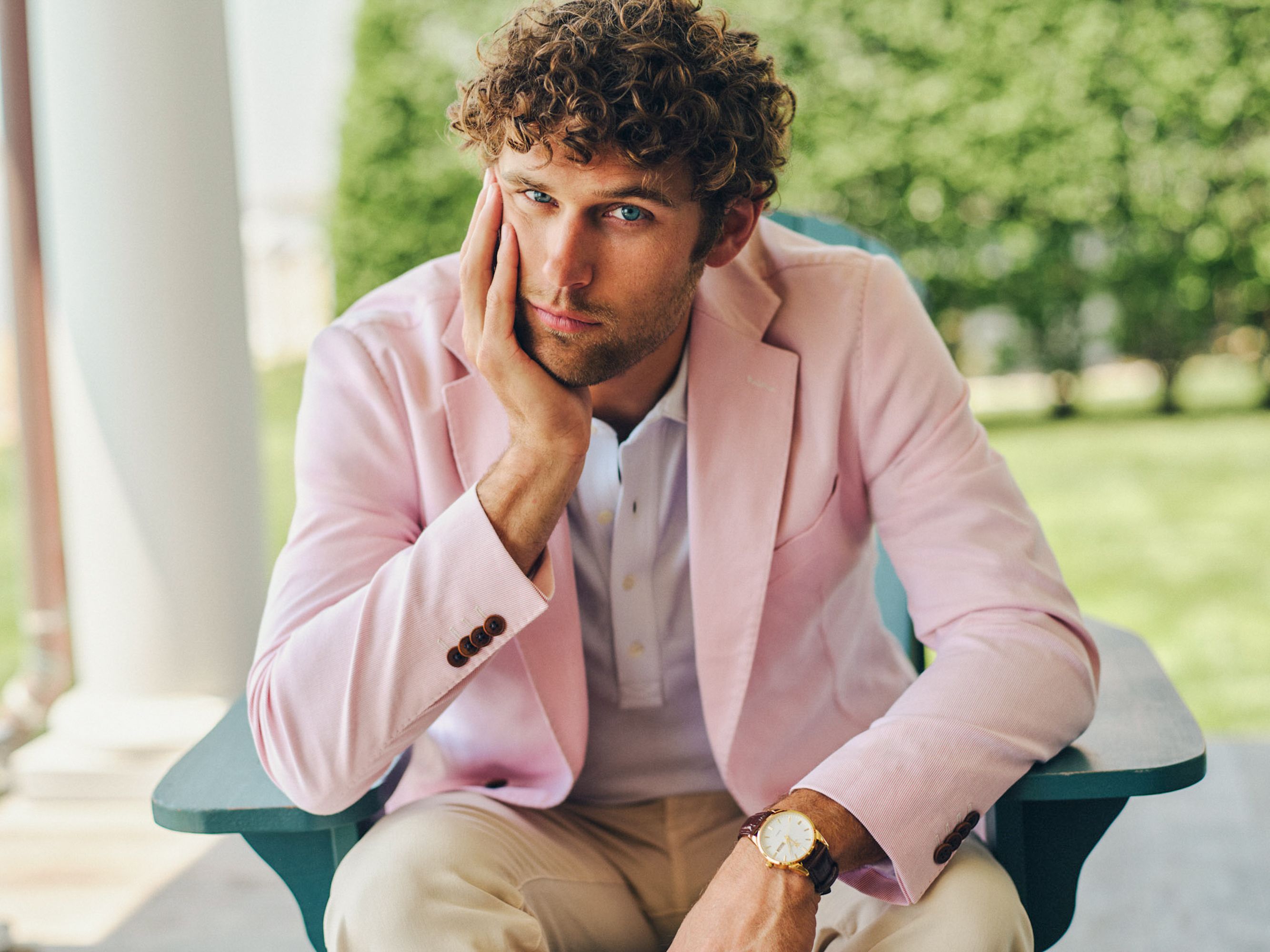 A man with curly hair and blue eyes, wearing a pink blazer, sits in a green outdoor chair, resting his chin on his hand and looking forward.