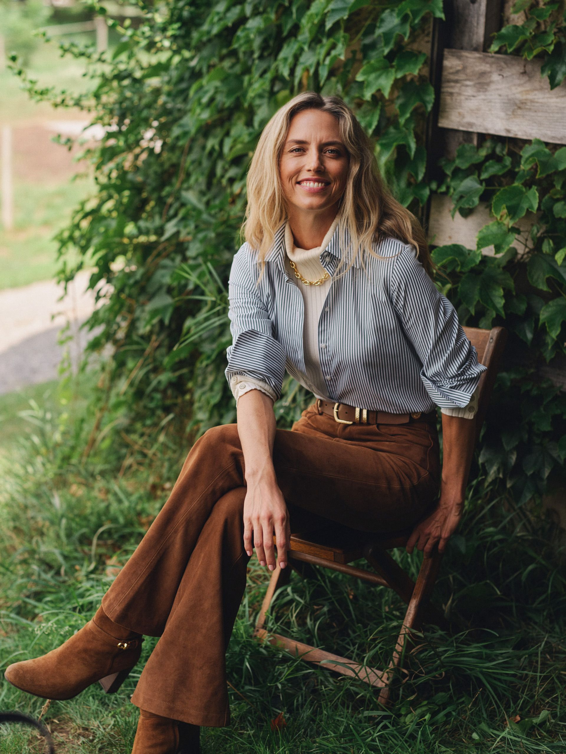 A smiling woman in a pinstripe shirt, turtleneck, brown suede pants, and boots sits on a wooden chair amidst green foliage.