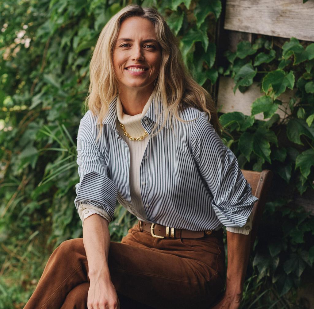 A smiling woman in a pinstripe shirt, turtleneck, brown suede pants, and boots sits on a wooden chair amidst green foliage.