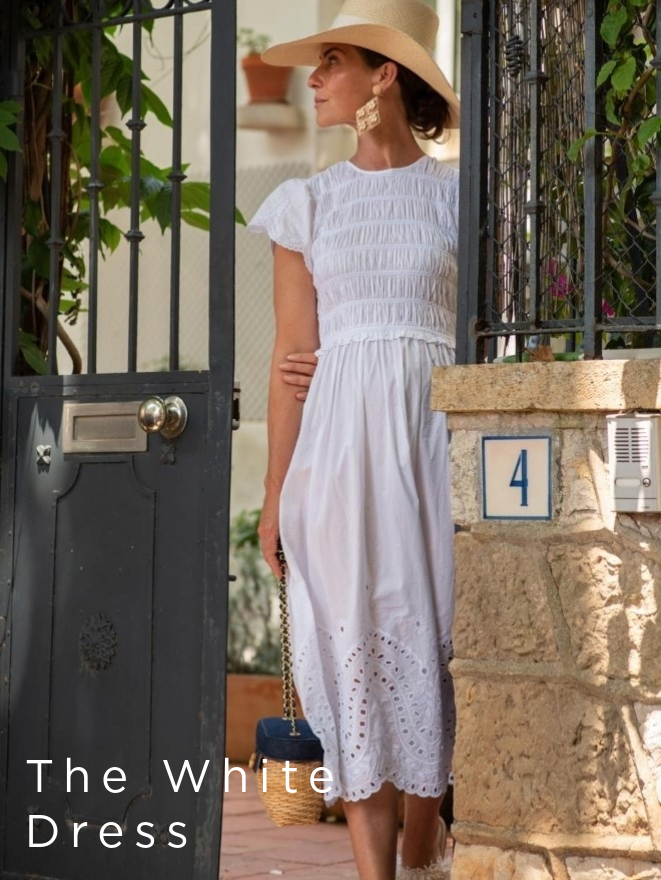 woman leaving gated doorway, wearing white midi dress with eyelet trim at sleeves and hem as well as a straw hat