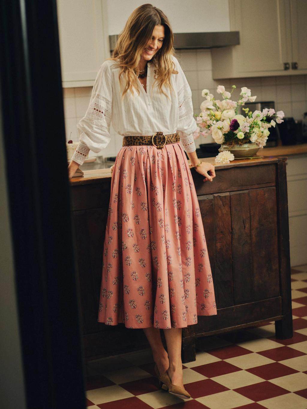 A smiling woman in a white blouse, leopard print belt, and pink floral skirt stands in a kitchen.