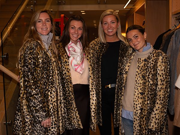 Four women standing together in a clothing store, smiling at the camera. From left to right: a woman in a leopard print coat with a striped scarf, a woman in a beige sweater with a pink and white patterned scarf and black pants, a woman in a leopard print coat over a black outfit, and a woman in a leopard print coat over a beige top and jeans. Warm lighting and clothing displays are visible in the background.