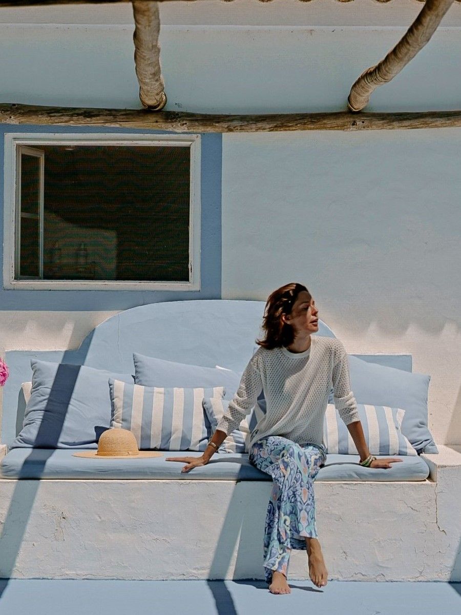 A woman in a white top and patterned blue pants sits barefoot on a blue and white outdoor bench with a straw hat.