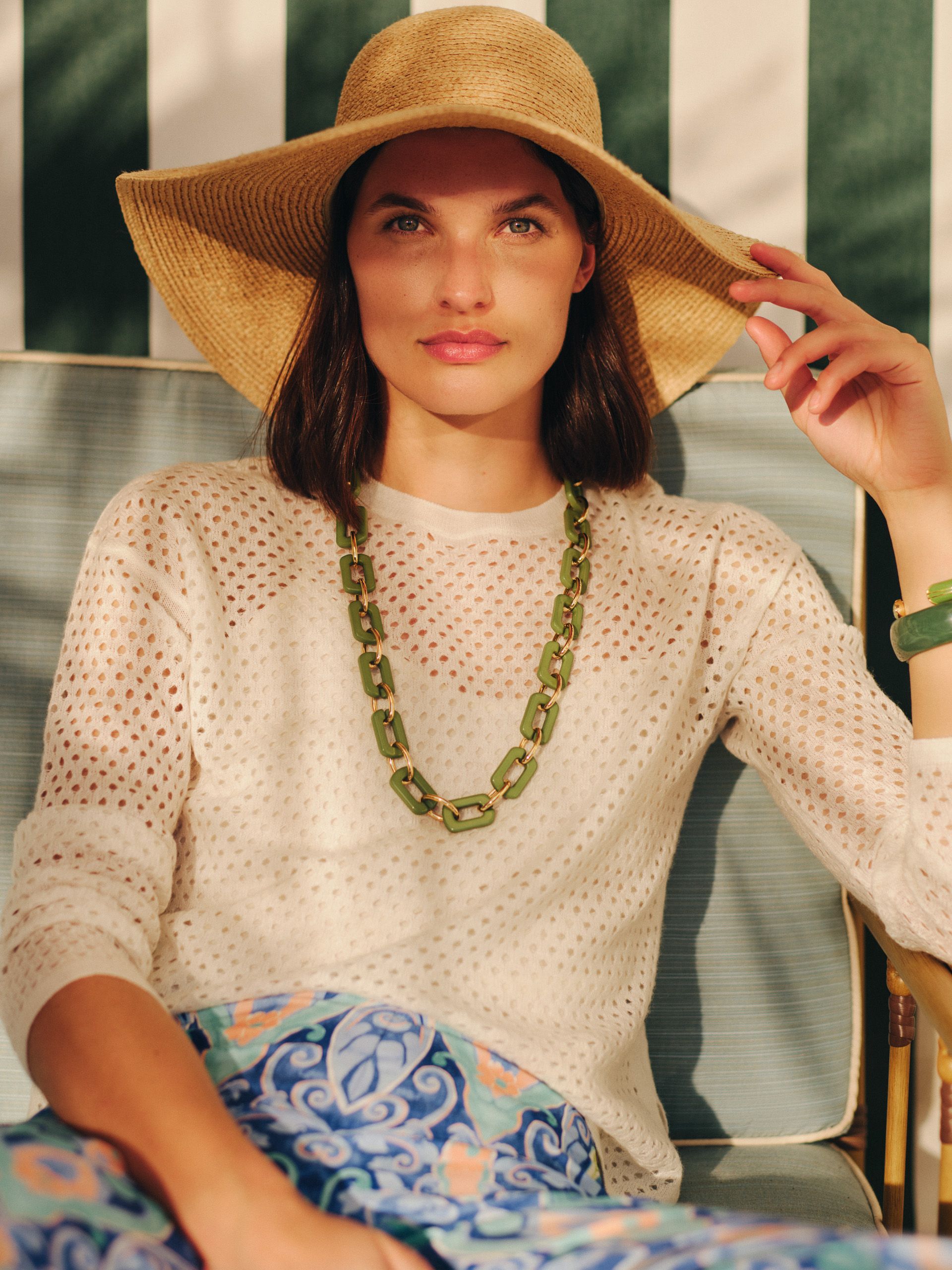 A woman in a straw hat, white open-knit sweater, green necklace, and blue patterned pants sits in a chair against a striped background.