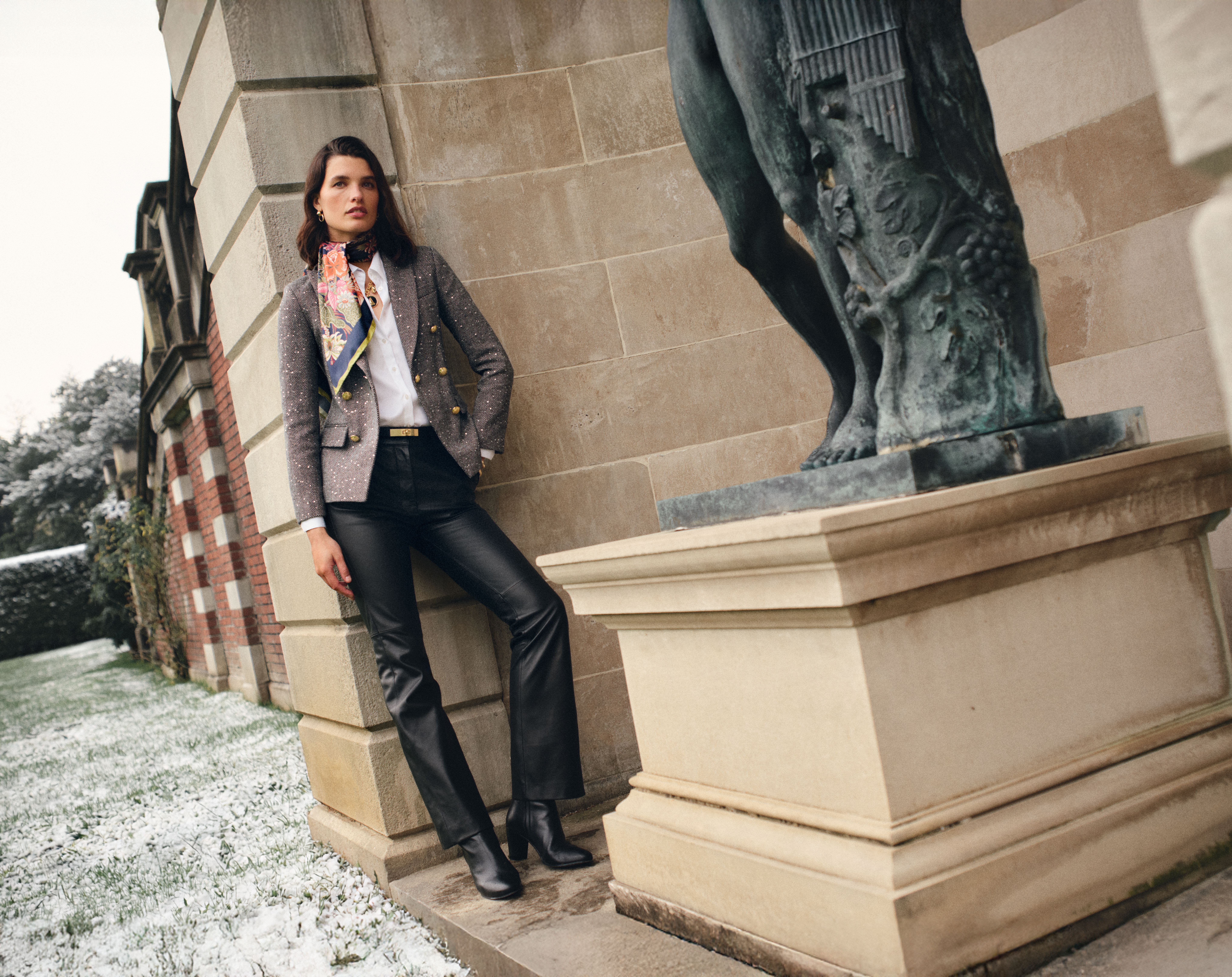 A woman in a grey blazer, colorful scarf, and black leather pants leans against a stone building with a statue, light snow on the ground.
