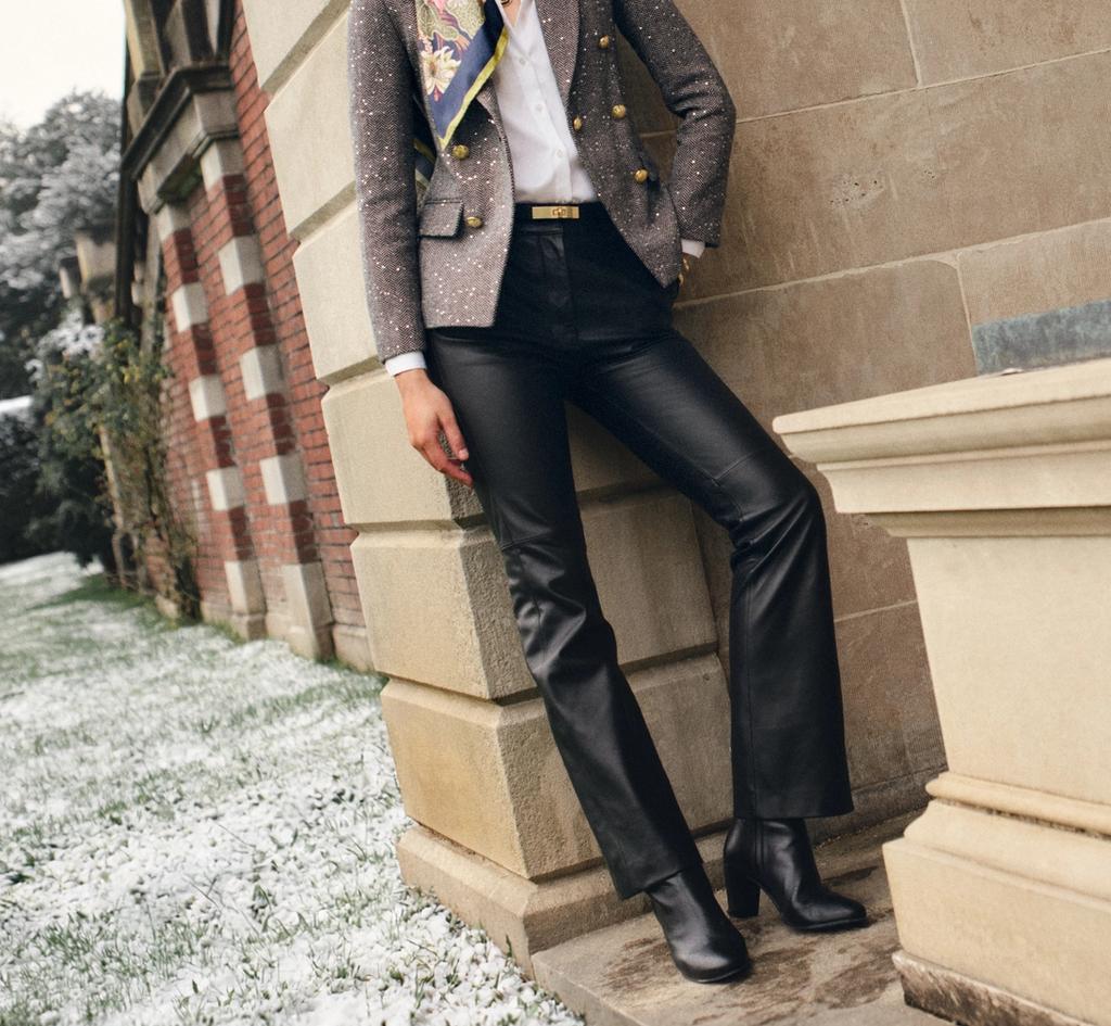 A woman in a grey blazer, colorful scarf, and black leather pants leans against a stone building with a statue, light snow on the ground.