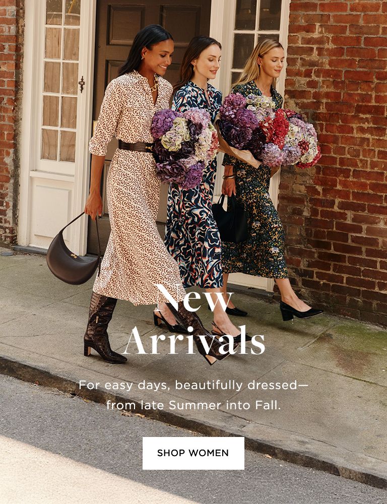 Three women standing together outdoors holding large bouquets of colorful flowers including purple, pink, red, and white blooms. They're wearing patterned dresses - one in a geometric print, one in an abstract blue and pink pattern, and one in a dark floral print. They're positioned in front of a brick building with white doors and windows. Text overlay reads "New Arrivals" and "For easy days, beautifully dressed—from late Summer into Fall" with a "SHOP WOMEN" button.