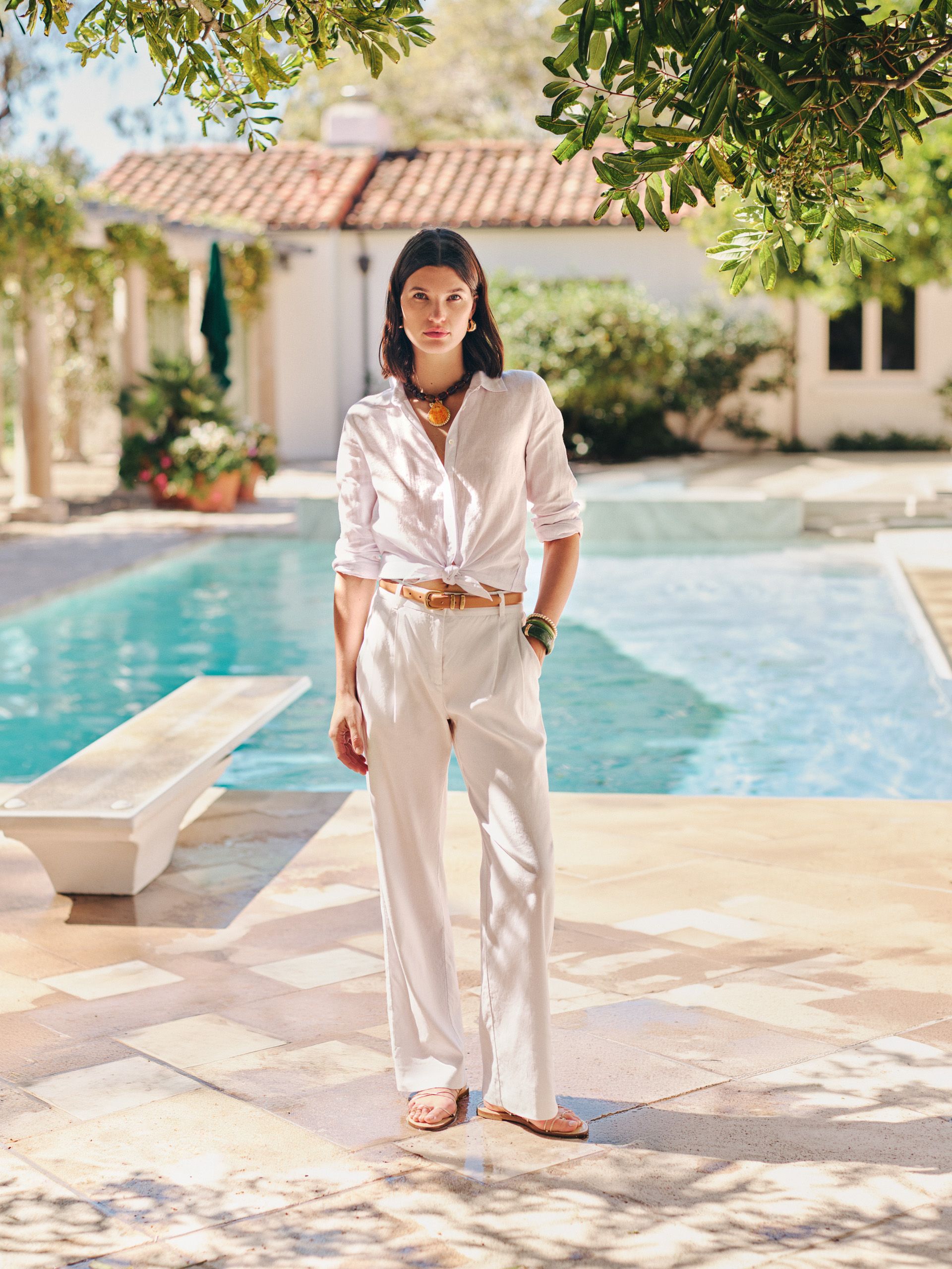 A woman in a white knotted shirt and white pants stands by a swimming pool with a Mediterranean-style house in the background.