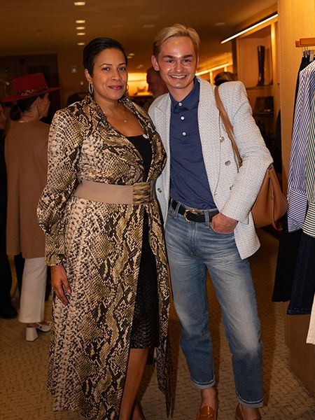 Two people posing together at an indoor event. On the left, a woman wearing a snake-print duster coat with a tan belt over a black dress. On the right, a person in a white textured blazer over a navy blue polo shirt and light blue jeans with rolled cuffs. Warm lighting and retail displays are visible in the background.