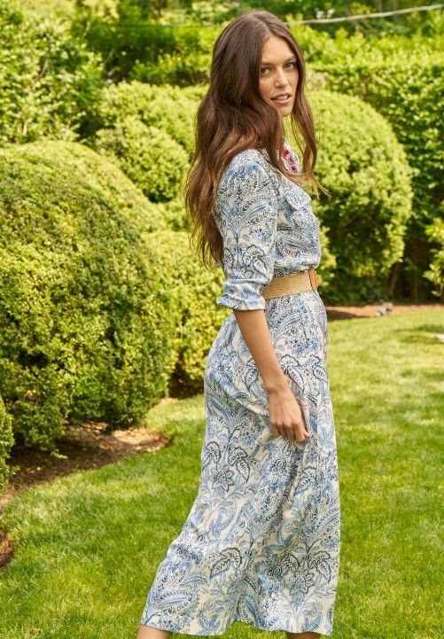 woman walking away in a lush green yard during the day, wearing a light blue paisley print silk midi shirtdress