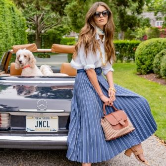 woman seated on back of vintage looking car in navy pleated skirt and white linen shirt