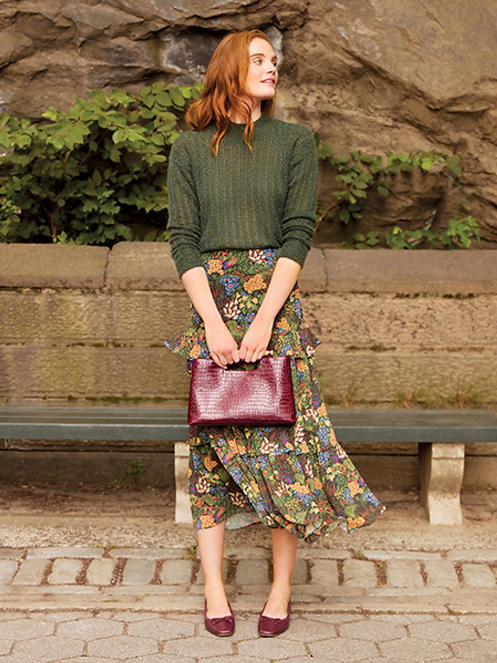 Women with red hair in green sweater and floral maxi skirt holding a maroon handbag.