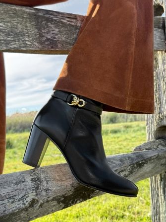 Black ankle boot with a gold buckle, worn with brown pants, resting on a wooden fence in a grassy field.