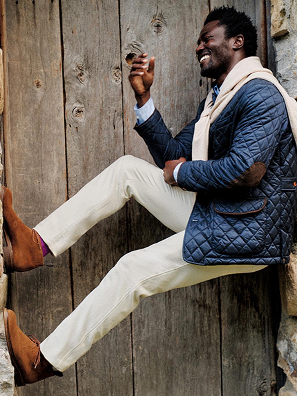 Smiling man resting against a wall wearing a navy blue puffer jacket, white pants, and brown suede boots.