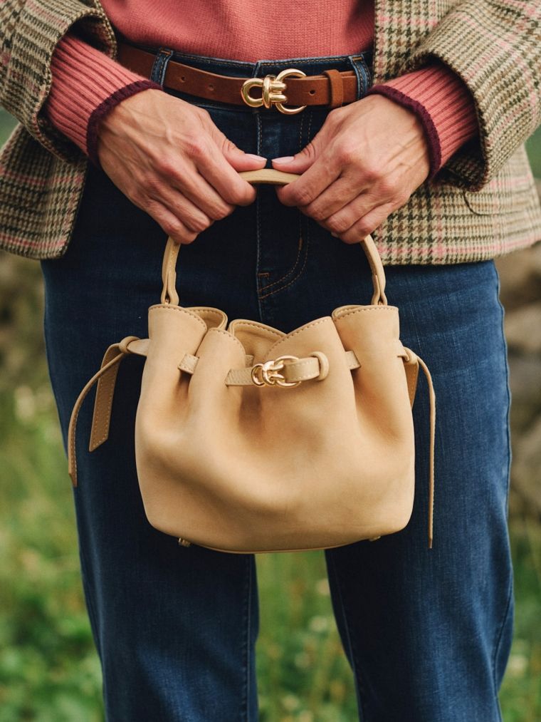 Person holding a tan bucket bag with a gold Gancini buckle, wearing a plaid jacket, rust sweater, and jeans.