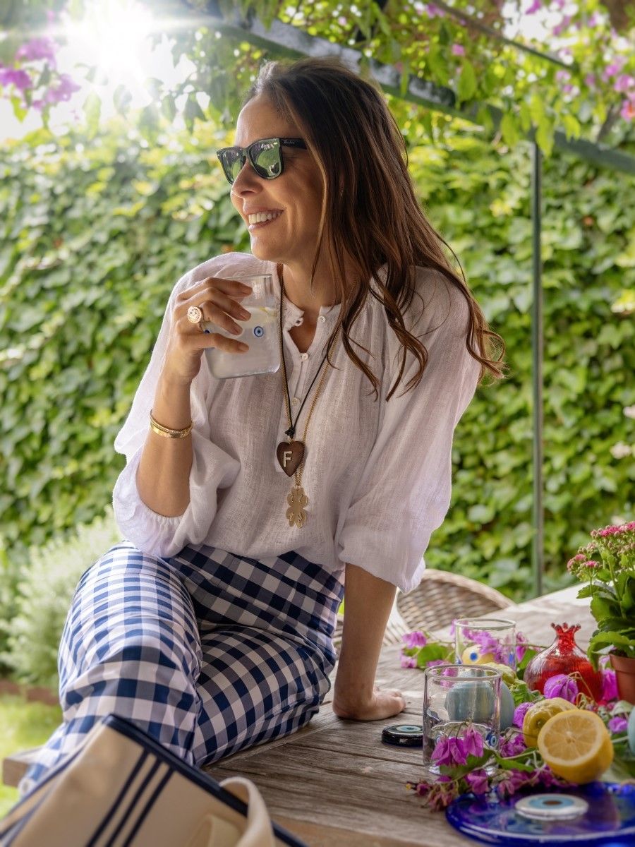A smiling woman in sunglasses, a white top, and blue gingham pants sits on an outdoor wooden table, holding a drink. Lemons, pomegranates, and flowers are also on the table.