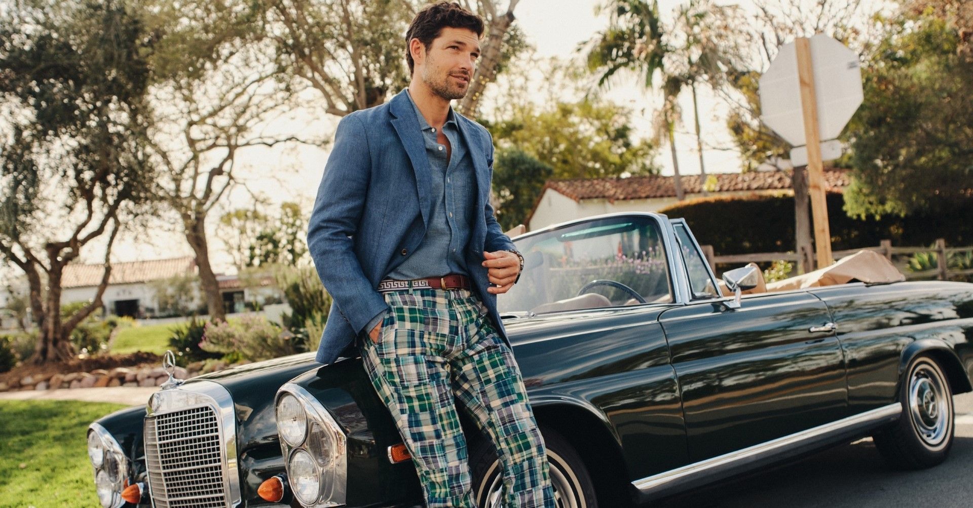 man outside leaning against a vintage looking convertible, wearing navy multicolor madras chino pants, a dark chambray linen blazer with matching shirt