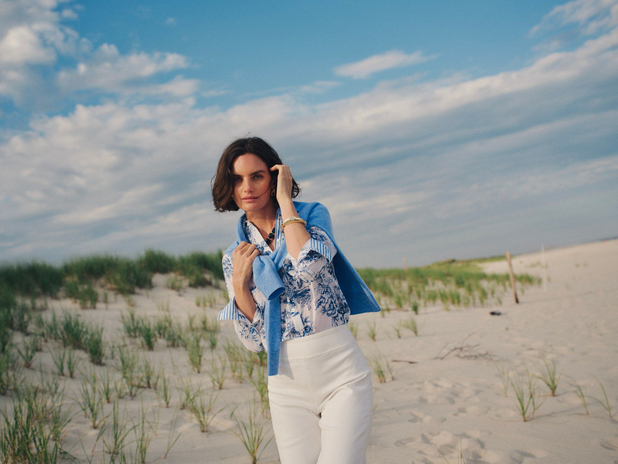 A woman in a blue floral top, white pants, and blue sweater on a sandy beach with dunes.