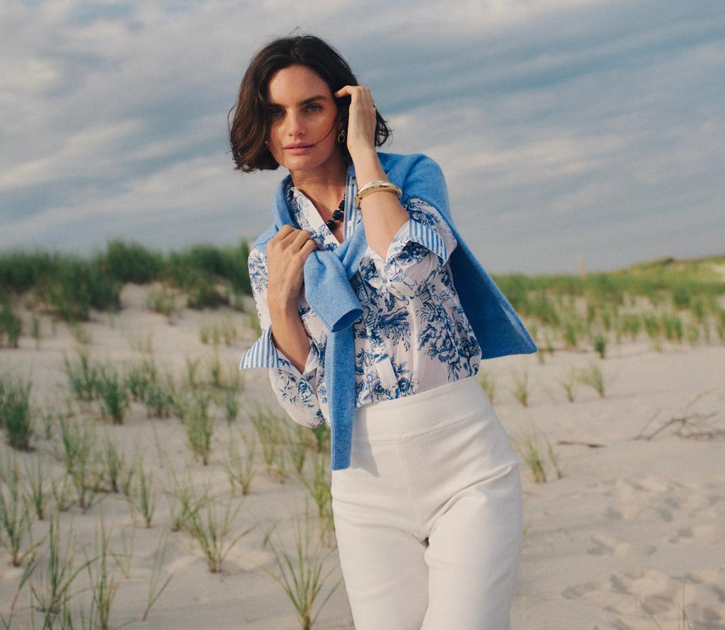 A woman in a blue floral shirt, white pants, and a blue sweater draped over her shoulders poses on a sandy beach with grassy dunes under a partly cloudy sky.