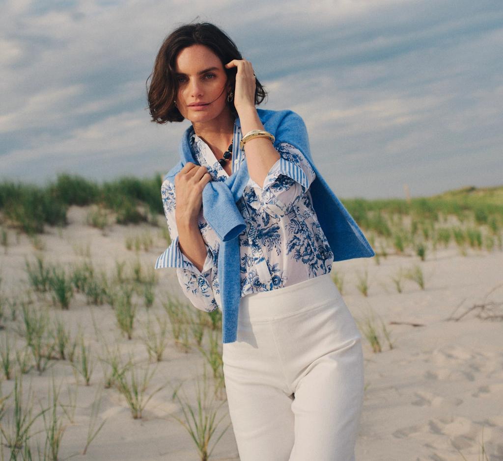 A woman in a blue floral top, white pants, and blue sweater on a sandy beach with dunes.