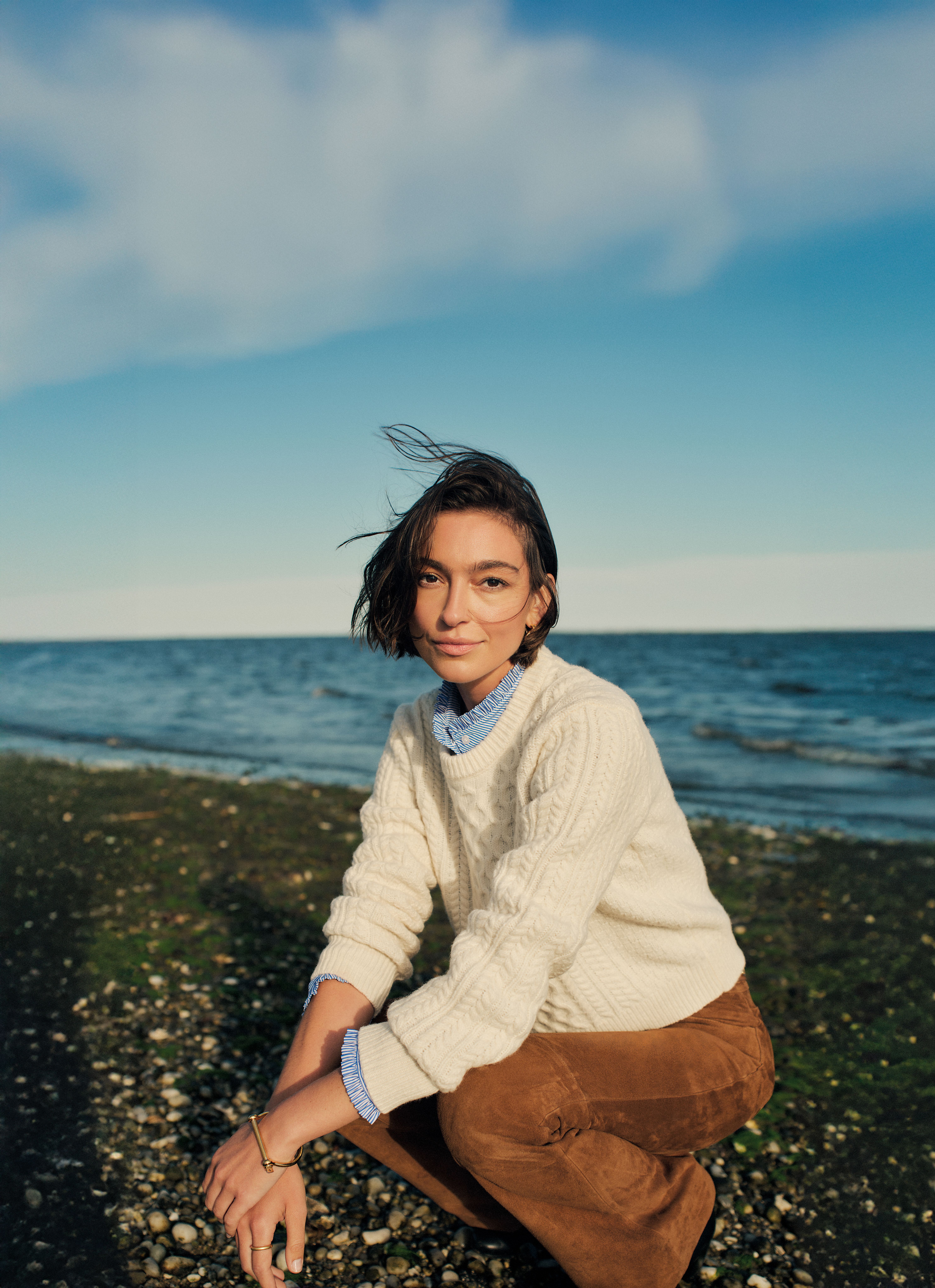 A woman with windblown hair crouches on a pebbly beach, wearing a cream cable knit sweater and brown pants, with the ocean and blue sky behind her.