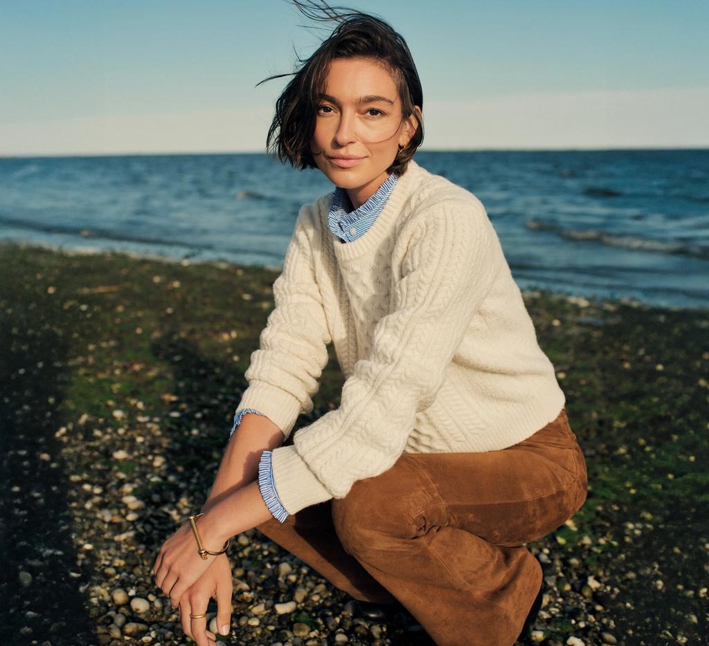 A woman in a cream cable-knit sweater and brown pants crouches on a pebbly beach with windblown hair, looking at the viewer.