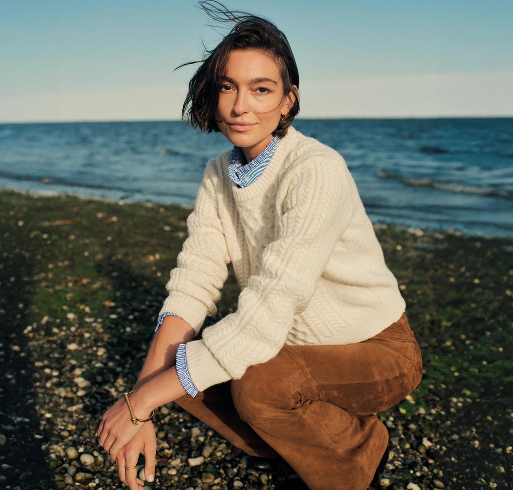 A woman with windblown hair crouches on a pebbly beach, wearing a cream cable knit sweater and brown pants, with the ocean and blue sky behind her.