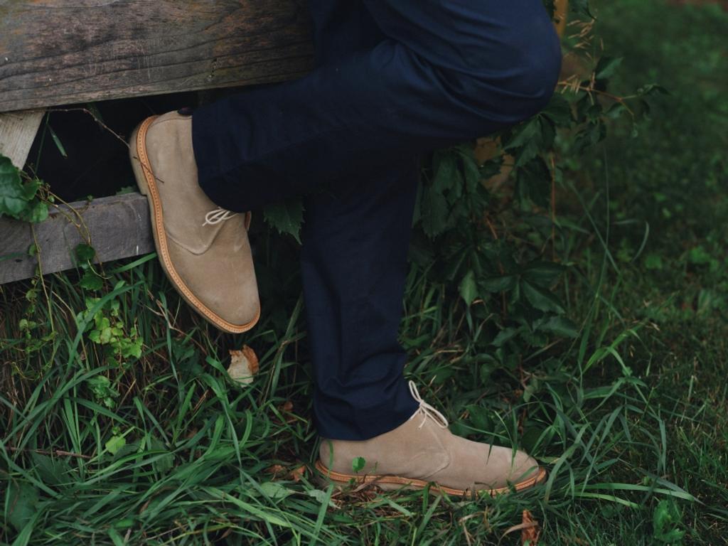 Person in navy pants and tan suede desert boots, standing in green grass with one foot resting on a wooden beam.