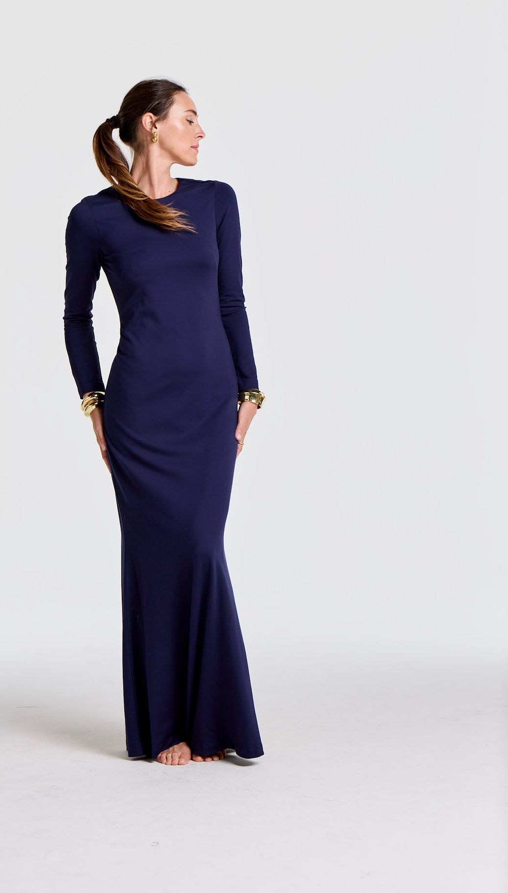 Barefoot woman in a navy blue long-sleeved maxi dress with gold cuffs and ponytail, looking right.