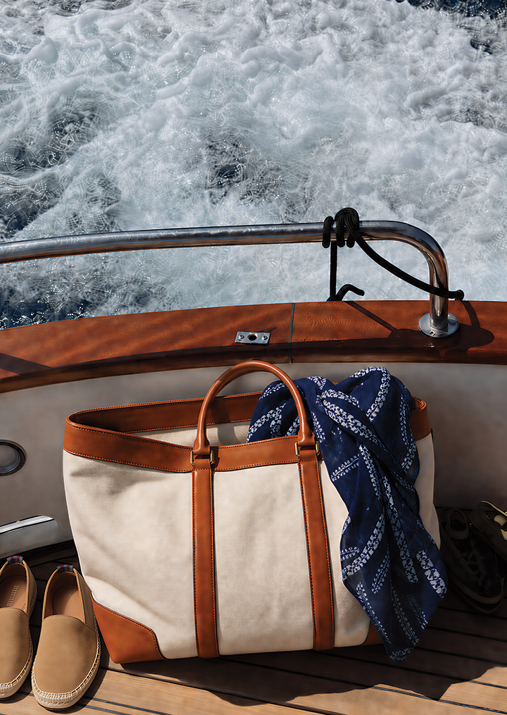 Cream and tan duffel bag, blue scarf, and tan espadrilles on a boat deck with foamy water behind.
