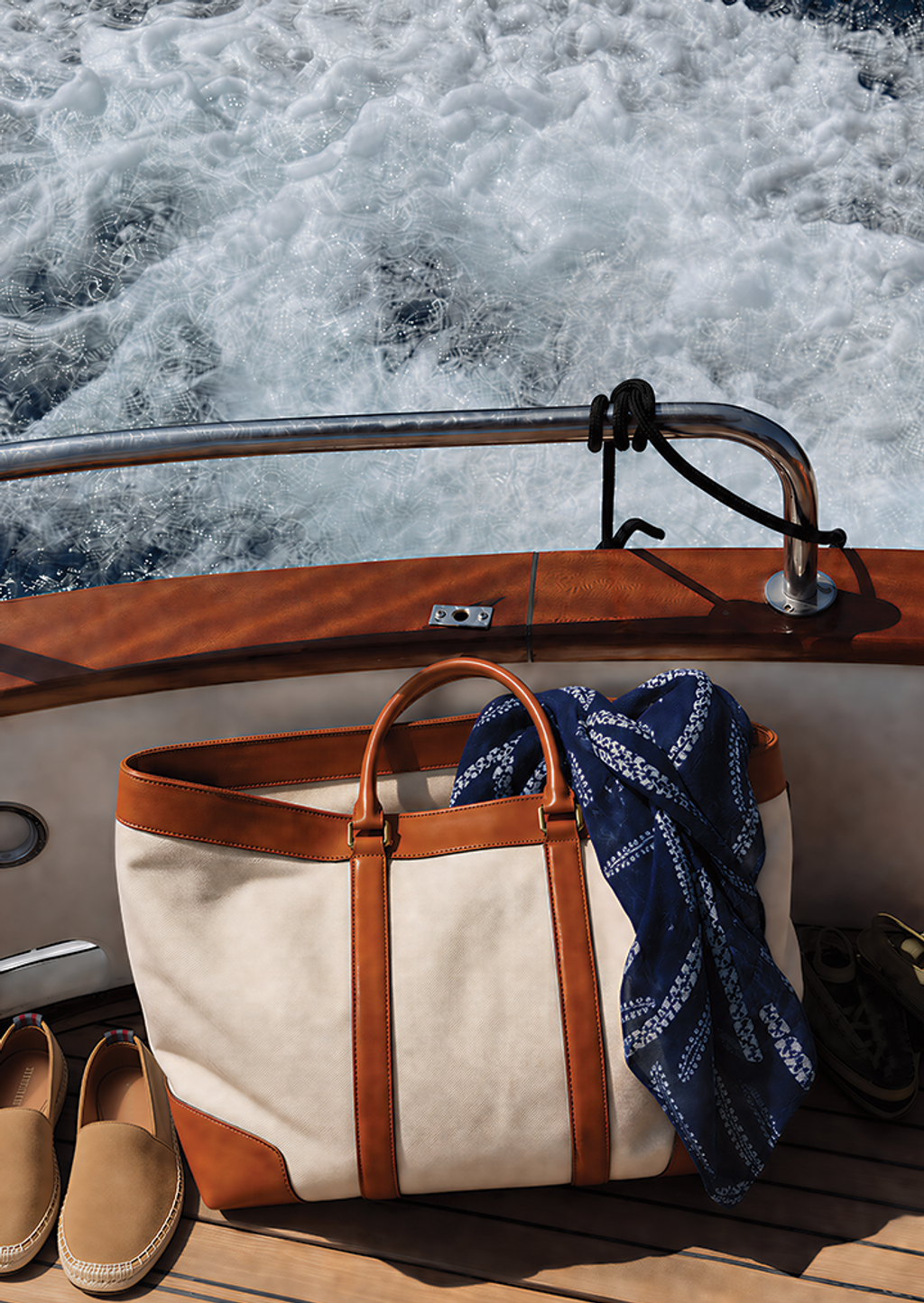 Cream and tan duffel bag, blue scarf, and tan espadrilles on a boat deck with foamy water behind.