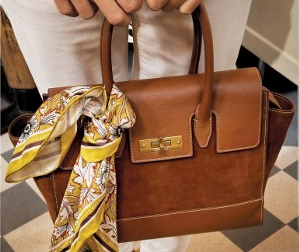 Brown leather and suede handbag with white stitching, a gold clasp, and a yellow patterned scarf tied to its handle, held by a person.