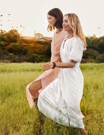 Two women in dresses walking through a sunny field at sunset.
